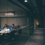 A group of people are sitting around a table with laptops and papers in a modern glass-walled office, engaged in a meeting. The hallway outside the meeting room is empty and dimly lit.