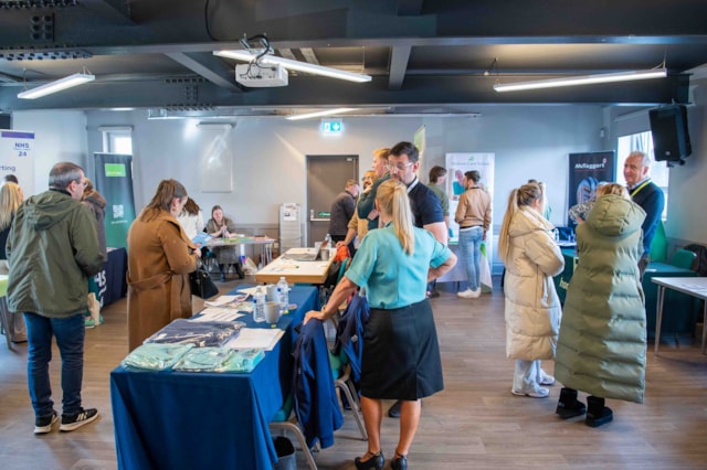People attend a job fair or information event indoors, standing and talking at tables with pamphlets and displays. The room is well-lit, and participants are casually dressed, with some wearing coats.