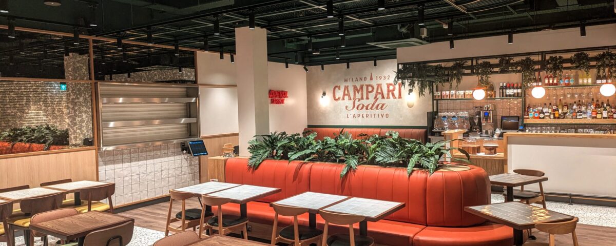 Modern restaurant interior with wooden tables and chairs, an orange curved booth with green plants, a bar area in the background, and a Campari Soda sign on the wall. The space is well-lit and neatly organized.