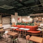 Modern restaurant interior with wooden tables and chairs, an orange curved booth with green plants, a bar area in the background, and a Campari Soda sign on the wall. The space is well-lit and neatly organized.