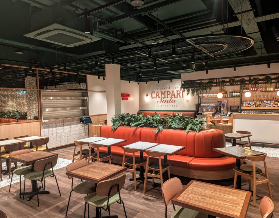 Modern restaurant interior with wooden tables and chairs, an orange curved booth with green plants, a bar area in the background, and a Campari Soda sign on the wall. The space is well-lit and neatly organized.