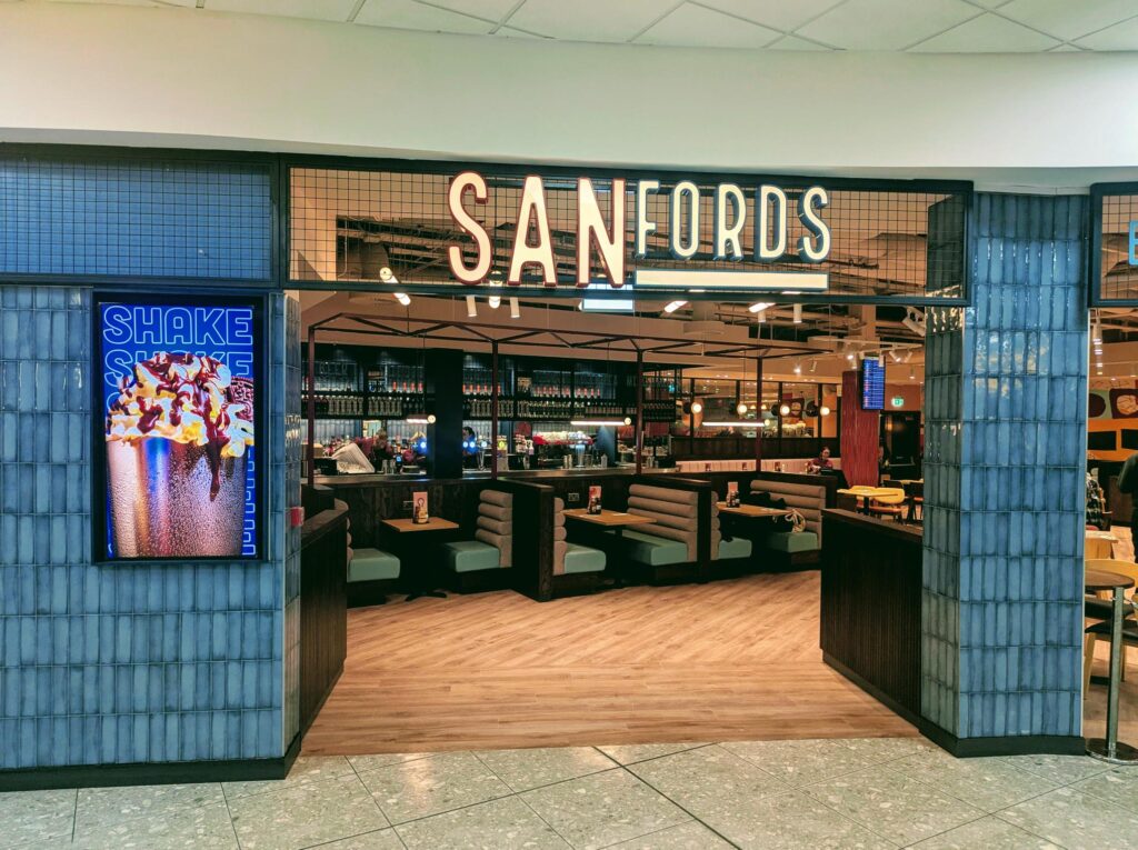 A restaurant entrance with a large "SANFORDS" sign above, featuring a modern interior with booth seating and warm lighting. A colorful "SHAKE" sign with images of milkshakes is displayed on the left side.
