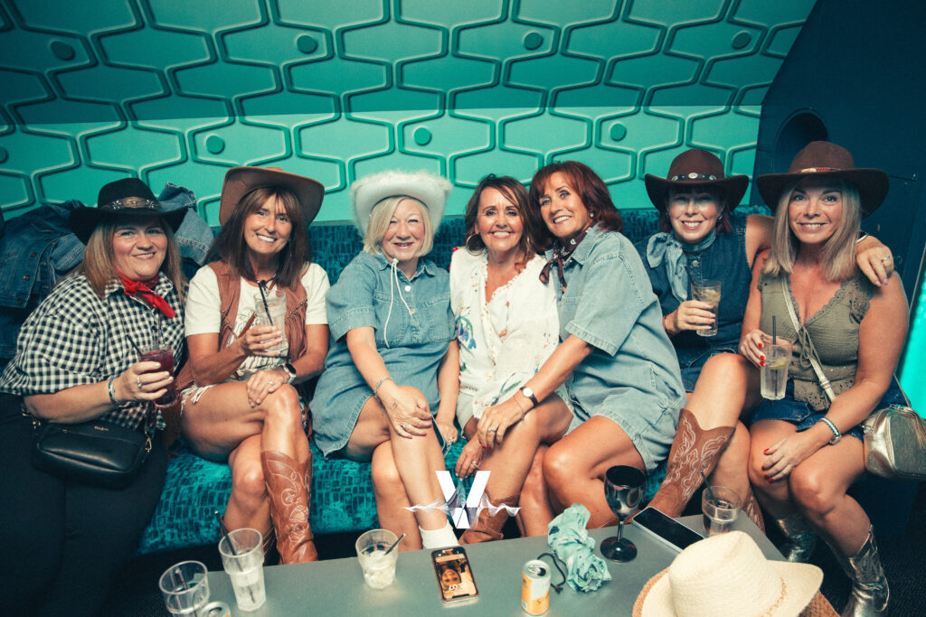 Six women dressed in cowboy hats and boots sit closely together on a blue couch, smiling and holding drinks in a room with a teal geometric wall. Empty cups and cans are on the table in front of them.