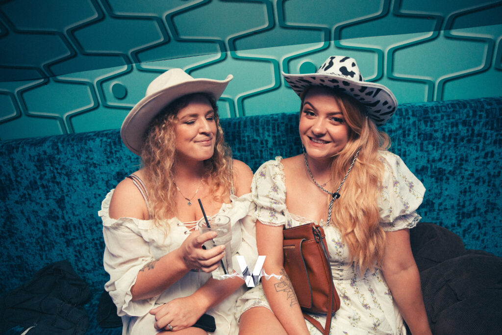 Two women in white dresses and cowboy hats sit on a blue couch in front of a patterned wall. One smiles at the camera while the other holds a drink and looks at her friend. Both appear relaxed and happy.