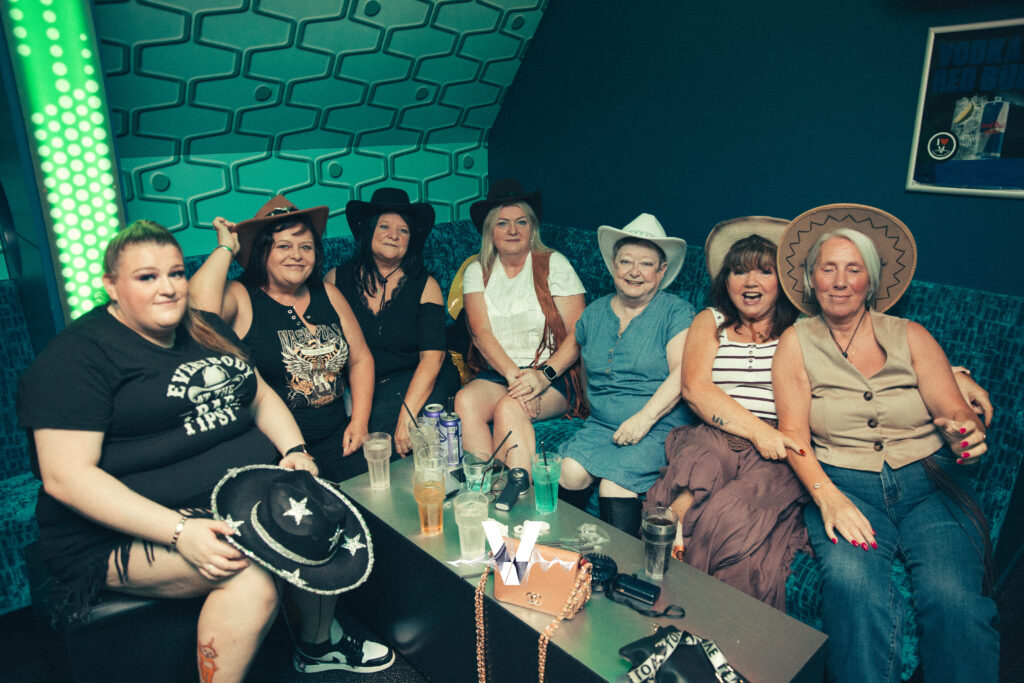 Seven women in cowboy hats and casual clothes sit together on a blue couch in a neon-lit room, smiling at the camera. Drinks and small bags are on a table in front of them. The atmosphere is festive and relaxed.