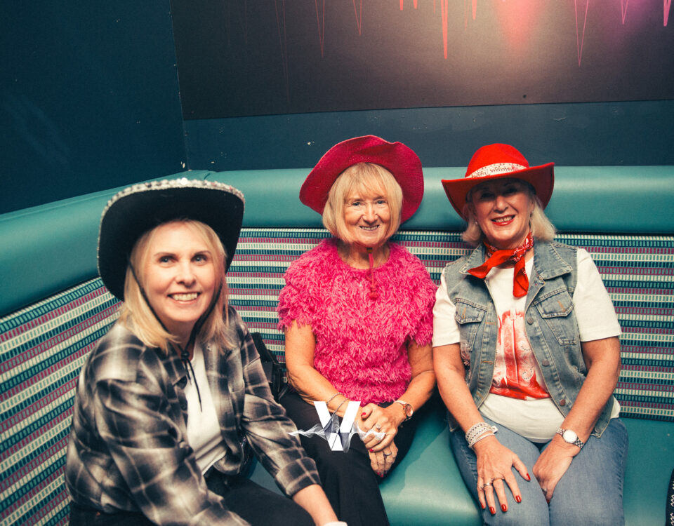Three smiling women sit together on a teal bench, dressed in Western-themed outfits with cowboy hats. One wears plaid, another a bright pink top, and the third has a red scarf and hat. The setting looks like a lounge or club.