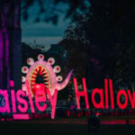 Large illuminated "Paisley Halloween" sign with a colorful monster tentacle and eyeballs decoration behind it, set outdoors at night with trees and historic buildings in the background.