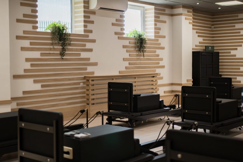 A modern Pilates studio with several black reformer machines, wooden slat wall accents, two windows with trailing green plants, and light streaming in from above.