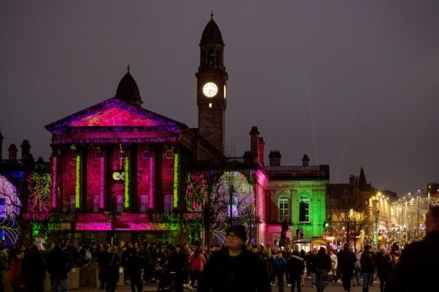 A large crowd gathers outside an old building illuminated by colorful light projections at night, with a clock tower in the background and city lights lining the street.