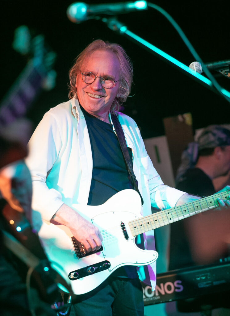 A smiling man with shoulder-length hair and glasses plays a white electric guitar on stage, wearing a white shirt over a black t-shirt. Microphones and another musician are visible in the background.