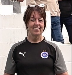 A woman with brown hair, wearing glasses on her head and a black and grey Puma sports shirt with a badge, stands smiling in front of white steps. People are visible in the background.