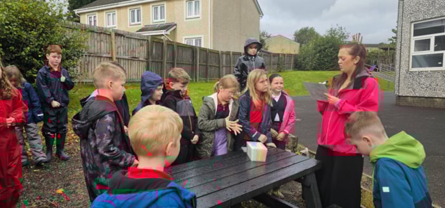 A group of school children and a woman stand and sit around a black picnic table outdoors. The woman holds a tablet and appears to be instructing or showing something to the children. Houses and a wooden fence are in the background.