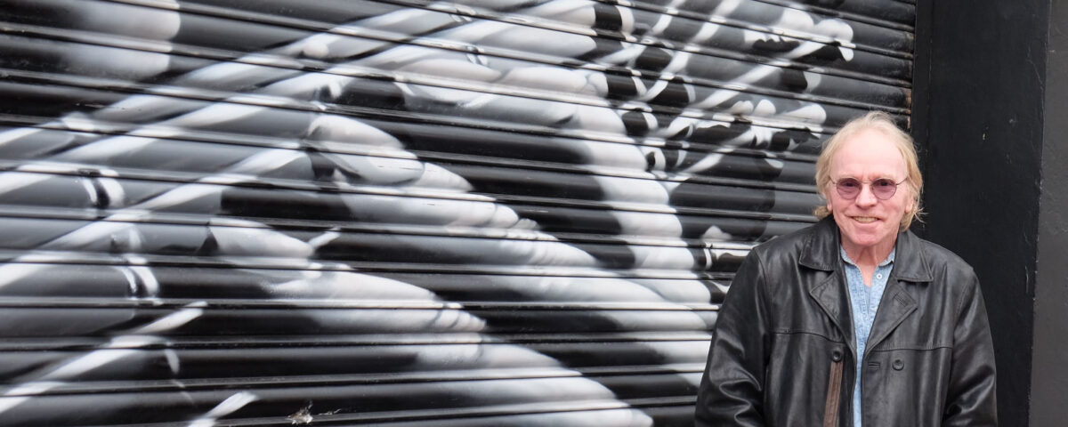 An older man in a black leather jacket and sunglasses stands in front of a mural on a metal shutter depicting a close-up of hands playing a guitar.