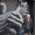 An older man in a black leather jacket and sunglasses stands in front of a mural on a metal shutter depicting a close-up of hands playing a guitar.