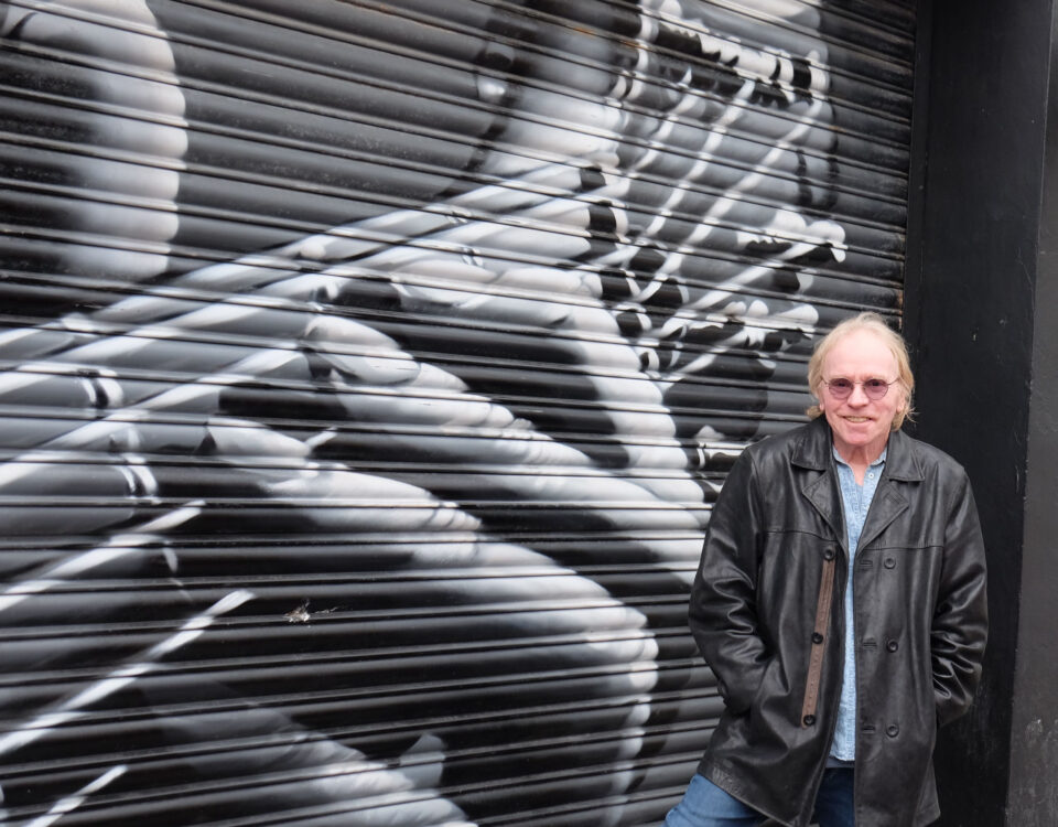 An older man in a black leather jacket and sunglasses stands in front of a mural on a metal shutter depicting a close-up of hands playing a guitar.