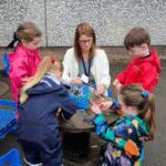 A woman and four children, all in colorful outdoor clothing, work together on a tech project at a round wooden table outside, with blue crates nearby and a green frog bin in the background.
