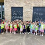 A group of young children in colorful clothes and neon safety vests stand in front of a brick wall with “Eva’s Day” written on two blackboards. The children are smiling and waving, celebrating together outdoors.