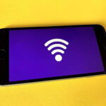 A smartphone with a purple screen displaying a white Wi-Fi symbol is placed on a bright yellow surface.