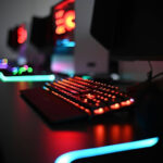 A close-up of a gaming keyboard and mouse glowing with colorful LED lights on a dark desk, with blurred computer monitors and more illuminated setups in the background.