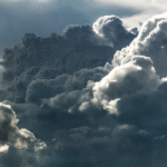 Large, dramatic clouds fill the sky, appearing dense and dark with sunlight peeking through in some areas, creating a contrast of bright and shadowed edges. The scene suggests an impending storm.