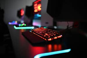 A close-up of a gaming keyboard and mouse glowing with colorful LED lights on a dark desk, with blurred computer monitors and more illuminated setups in the background.