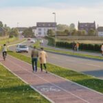 A paved road runs alongside a separate bike and pedestrian path. People walk, cycle, and drive on a sunny day, with houses, greenery, and blue sky in the background.