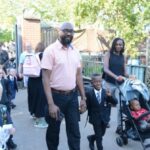 A group of children and adults, some in school uniforms, walk together outside on a sunny day. One man holds a boy's hand, while a woman pushes a stroller. Other families follow behind them.