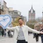 A smiling man stands on a busy city street with his arms open, holding a large heart-shaped "Love Local" sign. People walk in the background and a tall, historic building towers in the distance.