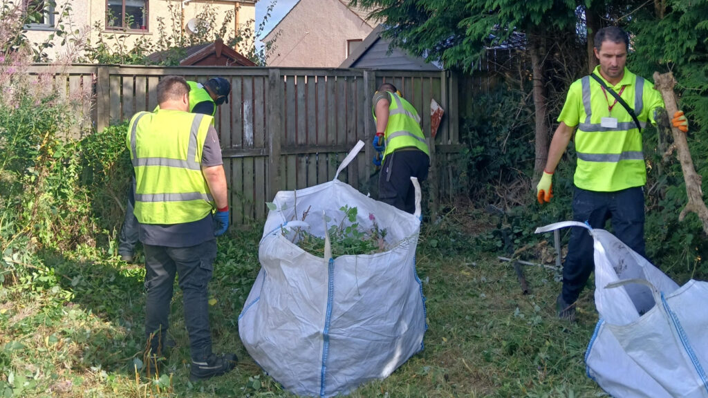 Four people in yellow safety vests work together in a garden, clearing overgrown plants and placing debris into a large white bag. A wooden fence and trees are in the background.
