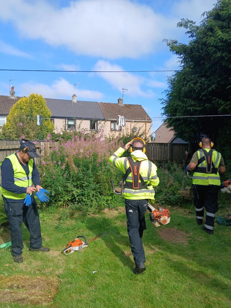 Three workers in high-visibility jackets and safety gear prepare equipment and put on gloves in a grassy yard, with houses and flowering bushes visible in the background.