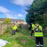 Four people wearing yellow safety vests clear overgrown plants and bushes in a garden with wooden fencing and houses in the background, under a partly cloudy blue sky.