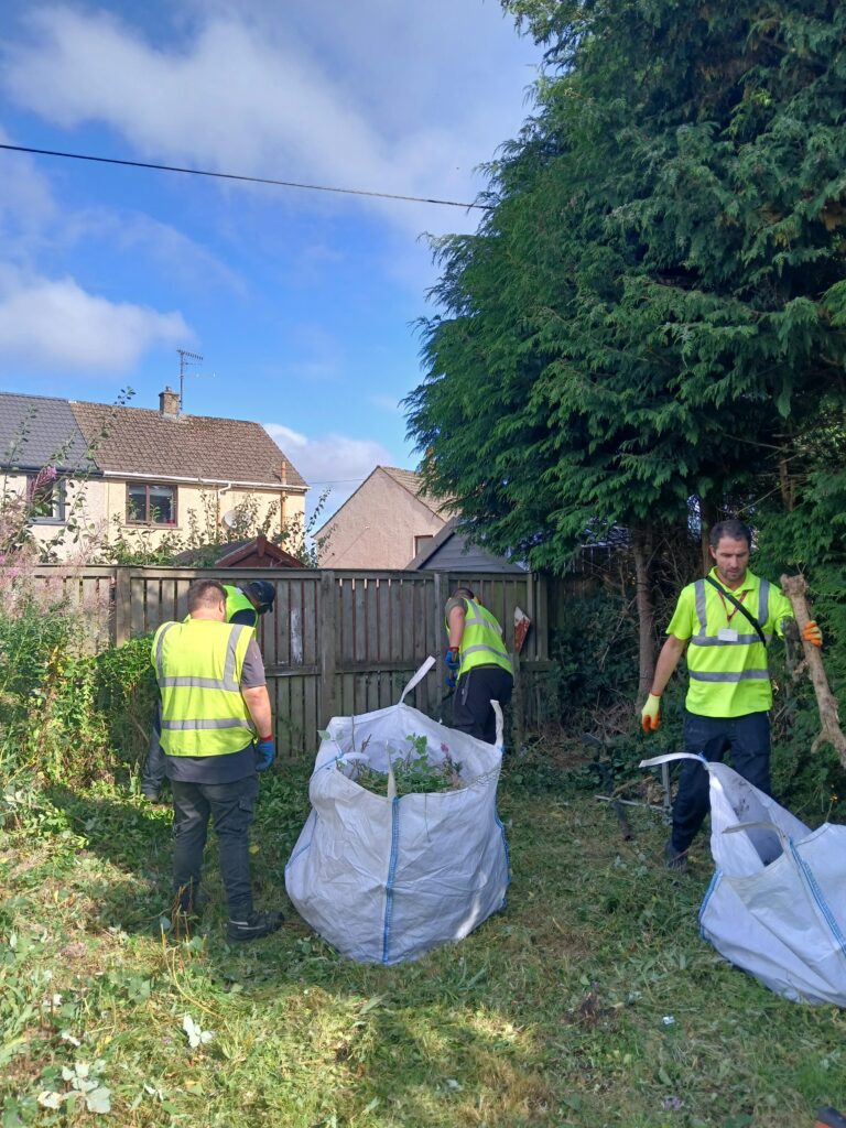 Four workers in high-visibility vests clear garden waste, placing it into large white bags in a grassy yard with tall trees and a wooden fence, under a partly cloudy sky.