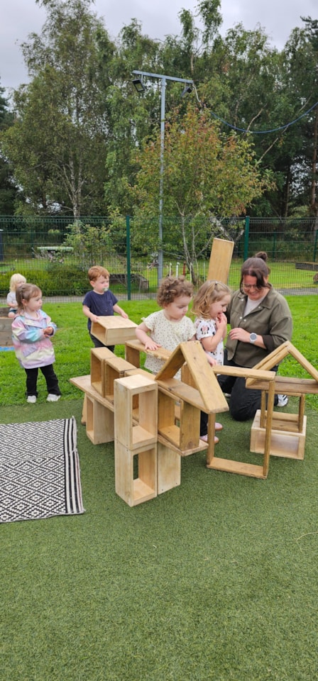 A group of young children and an adult play outdoors, building structures with large wooden blocks on green grass. Trees and a fence are visible in the background.