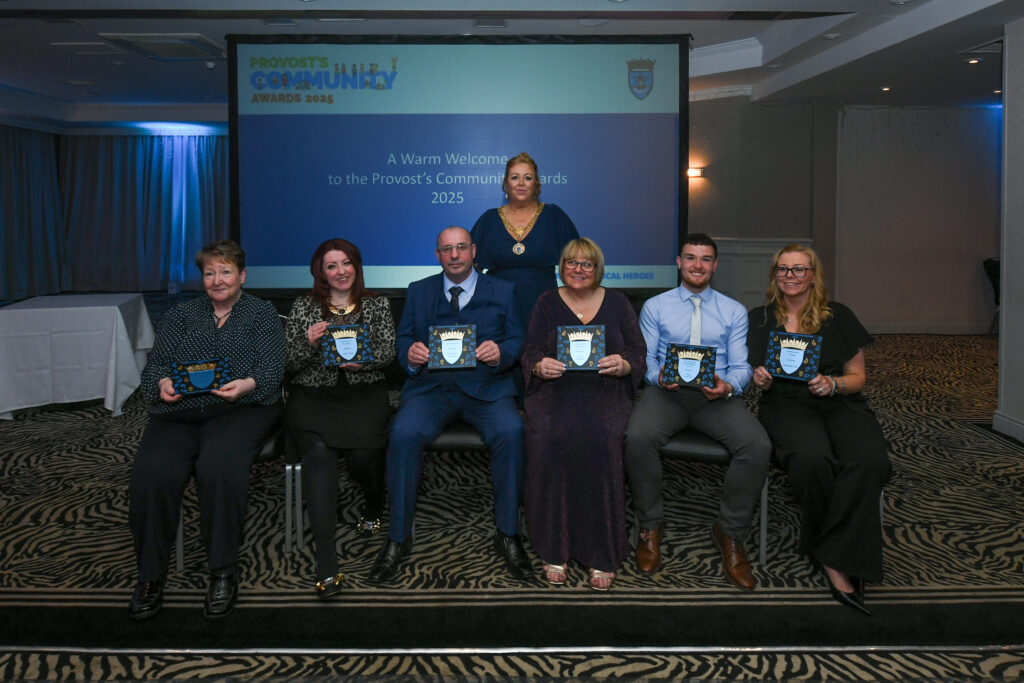 Seven people sit in a row holding award plaques, posing for a group photo at the Provost's Community Awards 2025 in a conference room. A woman stands behind them in front of a projection screen with event details.