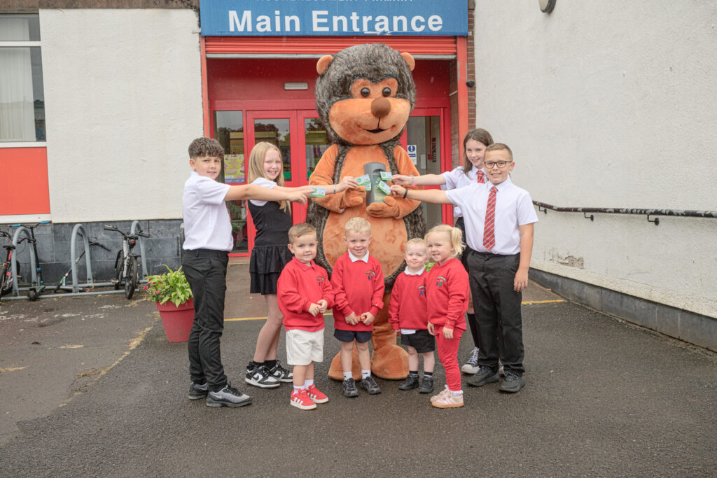 A group of children in school uniforms and a person in a large hedgehog mascot costume pose and smile outside a school’s main entrance, extending their arms to touch a large cup.