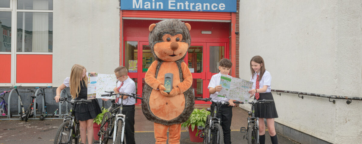 Four schoolchildren in uniforms with bicycles stand outside a school entrance, joined by a person in a large hedgehog costume holding a book. The school sign reads "Main Entrance Auchinlodment Primary".