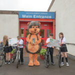 Four schoolchildren in uniforms with bicycles stand outside a school entrance, joined by a person in a large hedgehog costume holding a book. The school sign reads "Main Entrance Auchinlodment Primary".