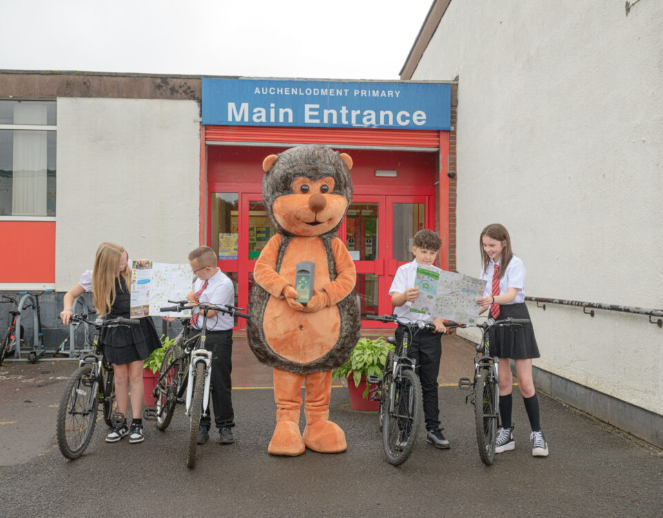 Four schoolchildren in uniforms with bicycles stand outside a school entrance, joined by a person in a large hedgehog costume holding a book. The school sign reads "Main Entrance Auchinlodment Primary".