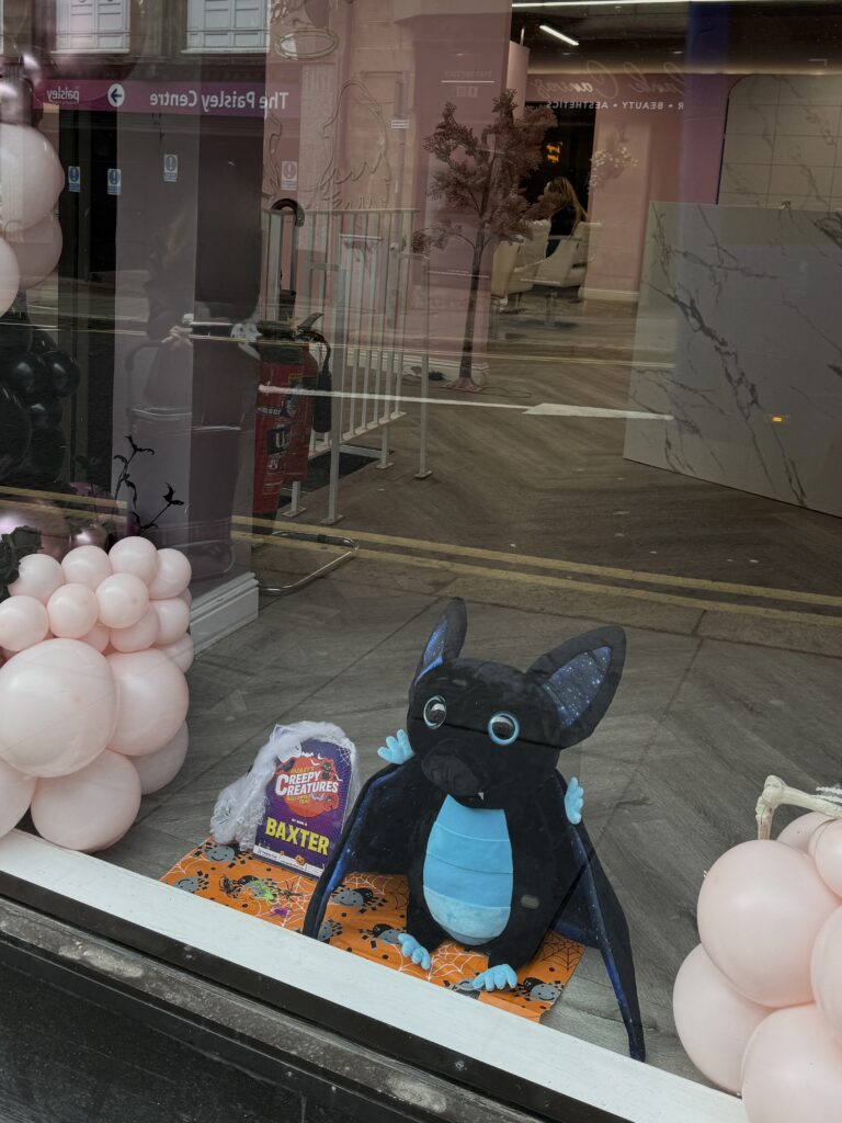 A plush bat toy with blue wings and belly sits in a store window, surrounded by pink balloons. A sign next to it reads "Baxter." Reflections of the street and buildings are visible in the glass.