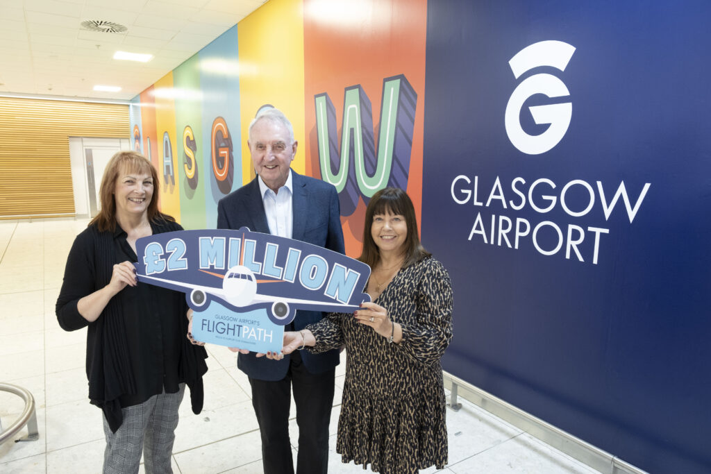 Three people stand smiling inside Glasgow Airport, holding a large sign shaped like a plane that reads "£2 Million, Glasgow Airport FlightPath," with a colorful Glasgow Airport wall sign in the background.
