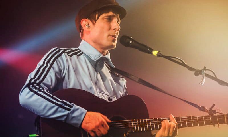 A musician wearing a cap and a light blue track jacket plays an acoustic guitar and sings into a microphone on stage, with colorful stage lights in the background.