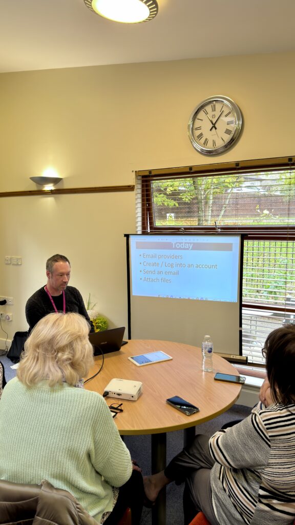 A man sits at a round table with three women, presenting a slideshow on a screen about email basics. A large clock and window are on the wall behind him, and various items are on the table.
