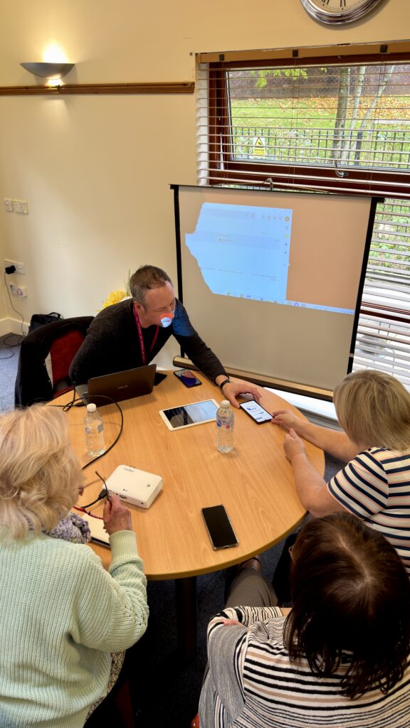 Four adults sit around a round table with laptops, smartphones, and water bottles, collaborating. One man is showing something on his phone to the group. A projector displays a screen on a nearby wall.