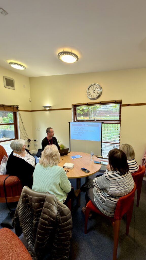 A group of five adults sits around a table, watching a presentation projected onto a screen in a cozy room with a clock and windows. The presenter uses a laptop, and notes are on the table.