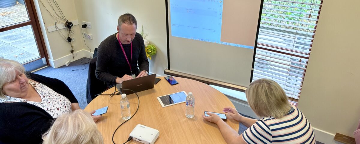 Five people sit around a circular table in a meeting room, some using mobile devices and laptops. A man leads the meeting with a projected screen behind him, and a large wall clock is visible above the window.