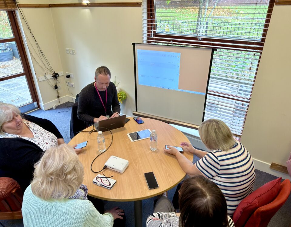 Five people sit around a circular table in a meeting room, some using mobile devices and laptops. A man leads the meeting with a projected screen behind him, and a large wall clock is visible above the window.