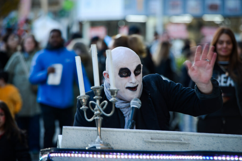 A person with white face makeup, dark eye circles, and bald head waves while speaking or singing into a microphone, standing behind a piano with a candelabra. A crowd of people is visible in the blurred background.