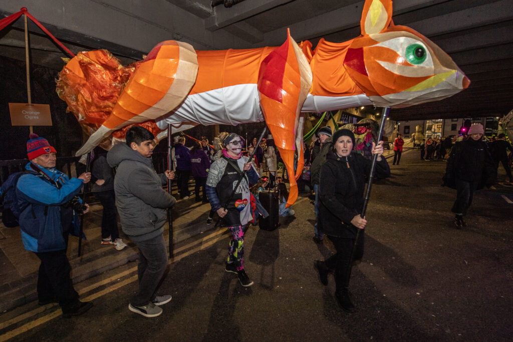 People carry a large, colorful, orange and white fox puppet under a bridge at night during a festive street parade. The group is dressed warmly, and some spectators watch in the background.