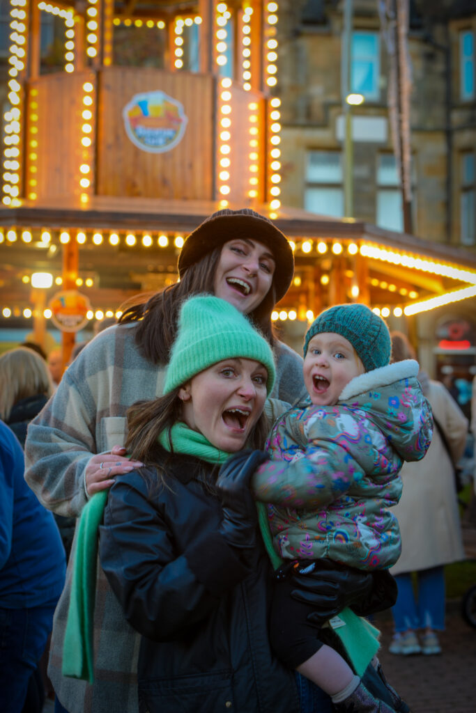 Three people, two adults and a child, stand smiling and laughing in front of a brightly lit carousel at an outdoor event. All are dressed warmly in hats and jackets. The background is bustling with people and lights.