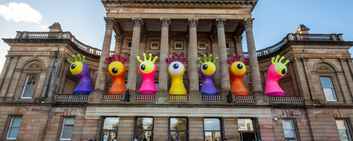 A historic building with tall columns displays large, colorful inflatable monster sculptures with big eyes and raised hands on its balcony. People walk and sit on the steps below under a clear, blue sky.